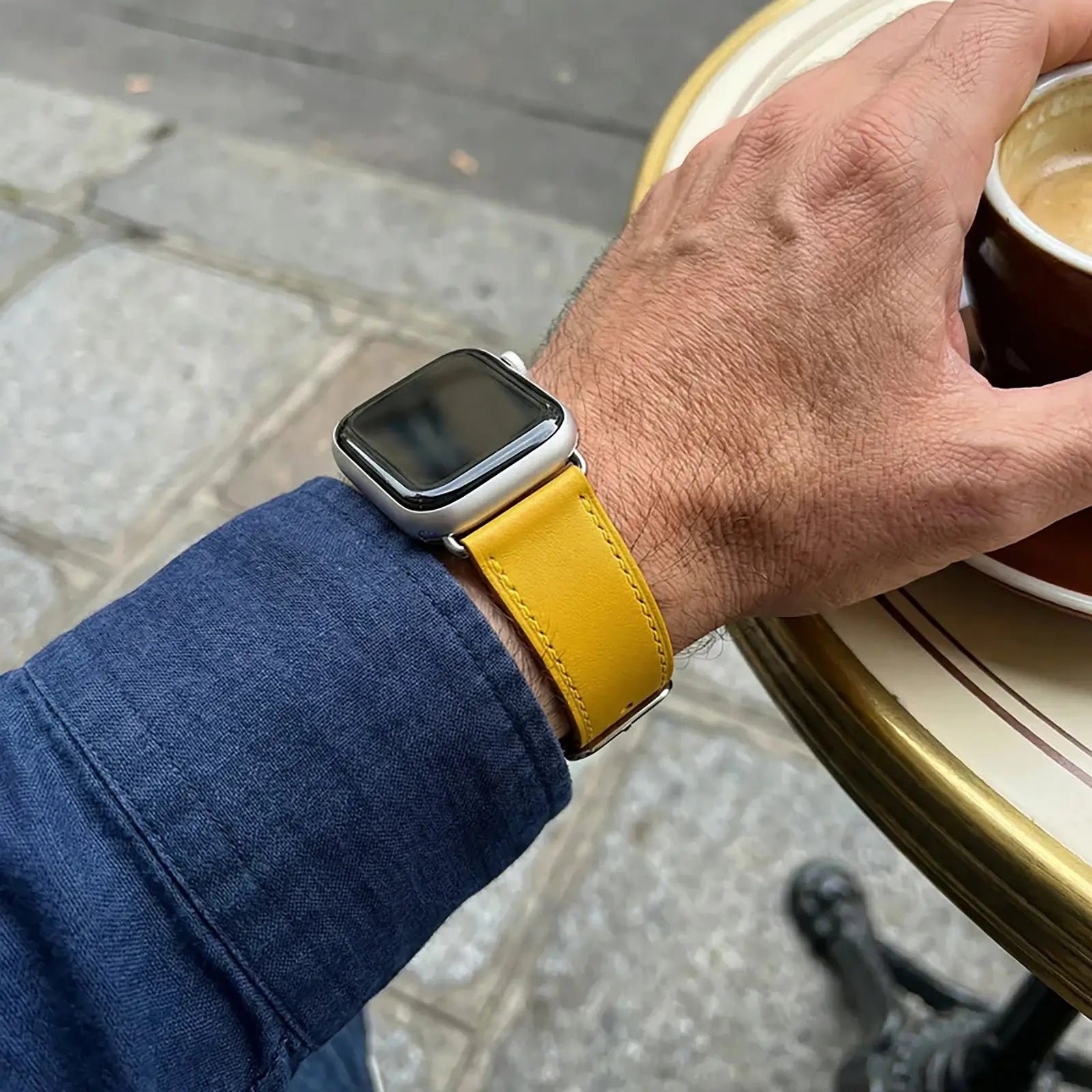 Hand wearing a smartwatch with a yellow strap, holding a cup of coffee on a sidewalk.
